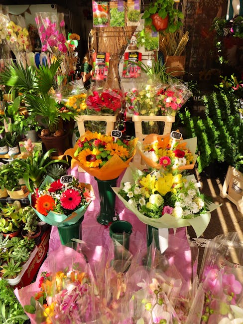A display of vibrant floral bouquets arranged on a pink table among various potted plants and greenery in a flower shop. The bouquets feature bright flowers such as red, yellow, pink, and white blooms