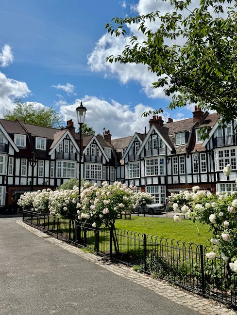 A row of traditional-style, black and white half-timbered terraced houses with steep, gabled roofs and numerous sash windows, set against a bright blue sky with scattered white clouds. In the foregrou