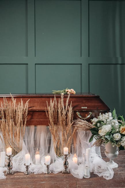 A floral arrangement displayed in front of a wooden coffin on a wooden floor, featuring tall, beige pampas grass and white flowers arranged in white vases. The arrangement is accented with small, lit 