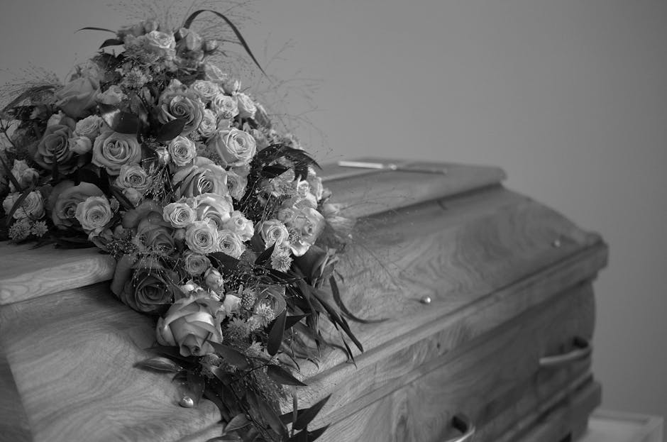 A floral arrangement of fresh funeral flowers resting atop a wooden casket, featuring a dense cluster of roses, carnations, and other blossoms with a mix of dark and light petals. The bouquet includes